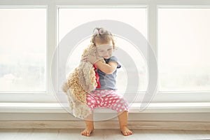 Adorable little girl hugging a teddy bear. Cute baby at home in white room is sitting near window.