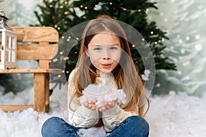 An adorable little girl in the interior of a Christmas studio blows artificial snow into the camera and rejoices at