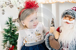 Adorable little girl looking at a large size Santa Claus doll with curiosity and fun surrounded by the joyful and festive room