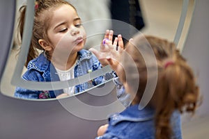 Adorable little girl making faces and looking at herself in the mirror, touching a button on a mirror, luminous mirror