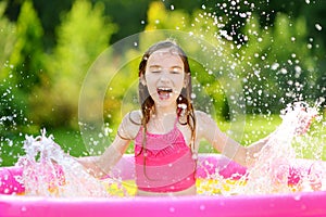 Adorable little girl playing in inflatable baby pool. Happy kid splashing in colorful garden play center on hot summer day.