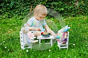 Adorable little girl playing with soft toys in park or garden and making them dinner with toy fruits and vegetables