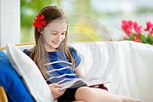 Adorable little girl reading a book in white living room on beautiful summer day.