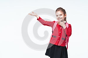 Adorable little girl in red school jacket, black dress, backpack pointing to empy space while posing on white studio