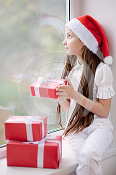 Pretty little girl in Santa hat dreaming by the window holding g
