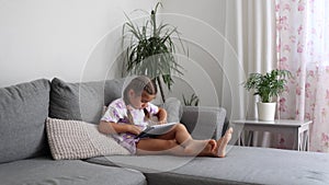 Adorable little girl sits at home on sofa and play games on tablet. Home mood
