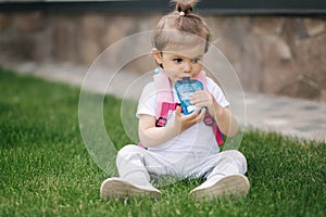 Adorable little girl sitting on the grass with backpack. Cute little girl drinking juise outdoors