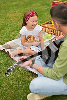 Adorable little girl sitting on a green grass in park and looking at her dad playing guitar