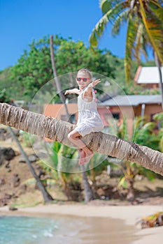 Adorable little girl sitting on palm tree during summer vacation on white beach