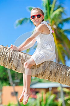 Adorable little girl sitting on palm tree during summer vacation on white beach