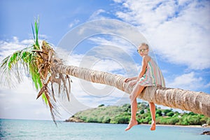 Adorable little girl sitting on palm tree during summer vacation on white beach