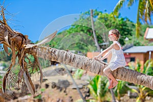 Adorable little girl sitting on palm tree during summer vacation on white beach