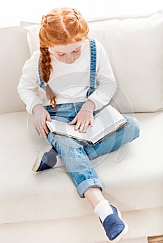 Adorable little girl sitting on sofa and reading book