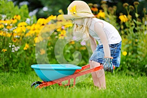 Adorable little girl in straw hat having fun with a toy wheelbarrow. Cute child playing farm outdoors.