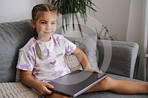 Adorable little girl take father& x27;s laptop and sits on sofa at home.