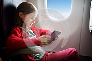 Adorable little girl traveling by an airplane. Child sitting by aircraft window and using a digital tablet