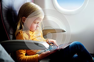 Adorable little girl traveling by an airplane. Child sitting by aircraft window and using a digital tablet