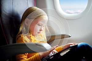 Adorable little girl traveling by an airplane. Child sitting by aircraft window and using a digital tablet