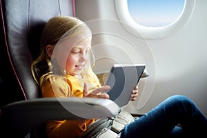 Adorable little girl traveling by an airplane. Child sitting by aircraft window and using a digital tablet