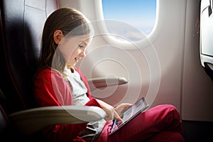 Adorable little girl traveling by an airplane. Child sitting by aircraft window and using a digital tablet