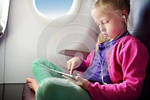 Adorable little girl traveling by an airplane. Child sitting by aircraft window and using a digital tablet during the flight.