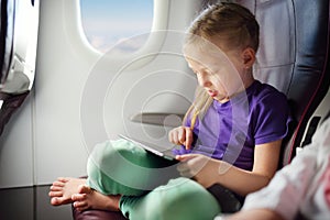 Adorable little girl traveling by an airplane. Child sitting by aircraft window and using a digital tablet during the flight.