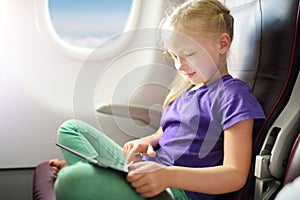 Adorable little girl traveling by an airplane. Child sitting by aircraft window and using a digital tablet during the flight.