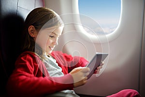 Adorable little girl traveling by an airplane. Child sitting by aircraft window and using a digital tablet during the flight.