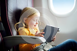 Adorable little girl traveling by an airplane. Child sitting by aircraft window and using a digital tablet during the flight.