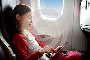 Adorable little girl traveling by an airplane. Child sitting by aircraft window and using a digital tablet during the flight.