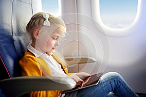 Adorable little girl traveling by an airplane. Child sitting by aircraft window and using a digital tablet during the flight. Trav