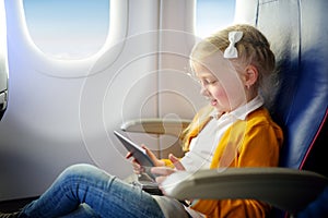 Adorable little girl traveling by an airplane. Child sitting by aircraft window and using a digital tablet during the flight. Trav