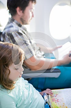 Adorable little girl traveling by an airplane with her father, sitting by aircraft window and reading books