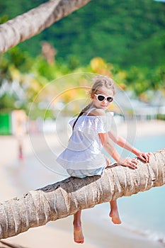 Adorable little girl at tropical beach sitting on palm tree during summer vacation