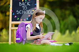 Adorable little girl using computer tablet while sitting on a grass on summer day