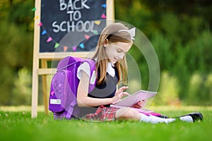 Adorable little girl using computer tablet while sitting on a grass on summer day