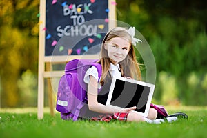 Adorable little girl using computer tablet while sitting on a grass on summer day