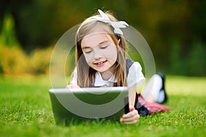 Adorable little girl using computer tablet while sitting on a grass on summer day