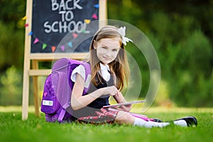 Adorable little girl using computer tablet while sitting on a grass on summer day