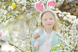 Adorable little girl wearing bunny ears in blooming cherry garden on beautiful spring day. Kid hanging Easter eggs on blossoming