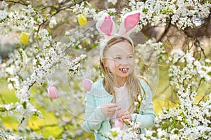 Adorable little girl wearing bunny ears in blooming cherry garden on beautiful spring day. Kid hanging Easter eggs on blossoming