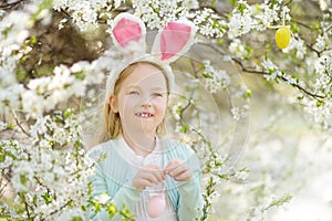 Adorable little girl wearing bunny ears in blooming cherry garden on beautiful spring day. Kid hanging Easter eggs on blossoming