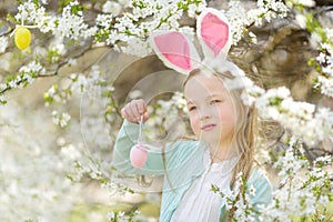 Adorable little girl wearing bunny ears in blooming cherry garden on beautiful spring day. Kid hanging Easter eggs on blossoming