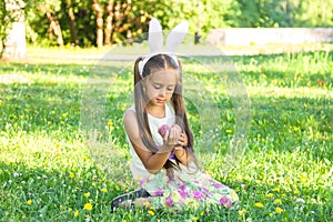 Adorable little girl wearing bunny ears on Easter day. Girl sitting on a grass and holding painted Easter eggs