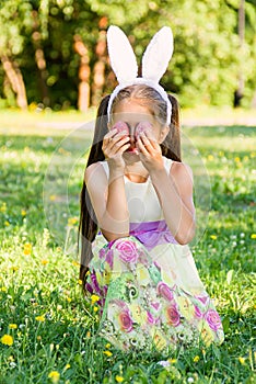 Adorable little girl wearing bunny ears on Easter day. Girl sitting on a grass and holding painted Easter eggs on her eyes,