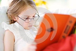 Adorable little girl wearing eyeglasses reading a book in white living room