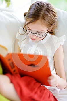 Adorable little girl wearing eyeglasses reading a book in white living room on summer day