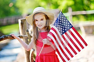 Adorable little girl wearing hat holding american flag outdoors on beautiful summer day