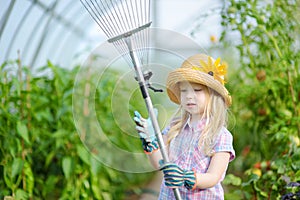 Adorable little girl wearing straw hat and childrens garden gloves playing with her toy garden tools in a greenhouse on summer day