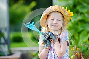 Adorable little girl wearing straw hat and childrens garden gloves playing with her toy garden tools in a greenhouse